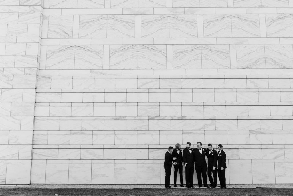 Groom and groomsmen stand against a white wall talking