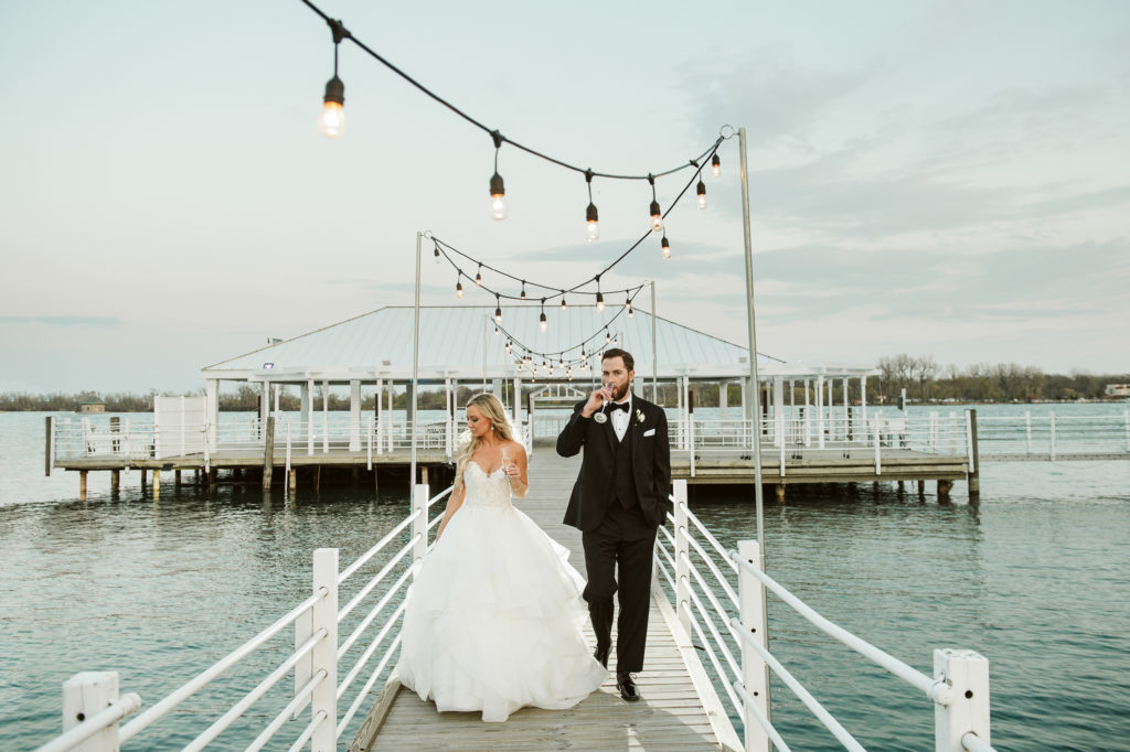 Bride and groom walk on dock while drinking champagne
