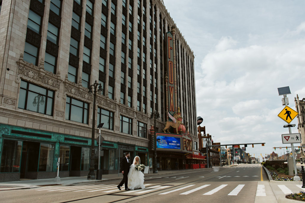 Bride and groom walk across a sunny street with Fox Theater behind them