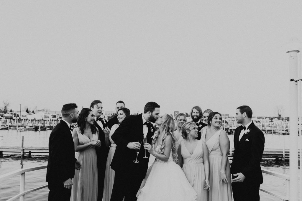Bride and groom hold champagne glasses and kiss in front of bridal party
